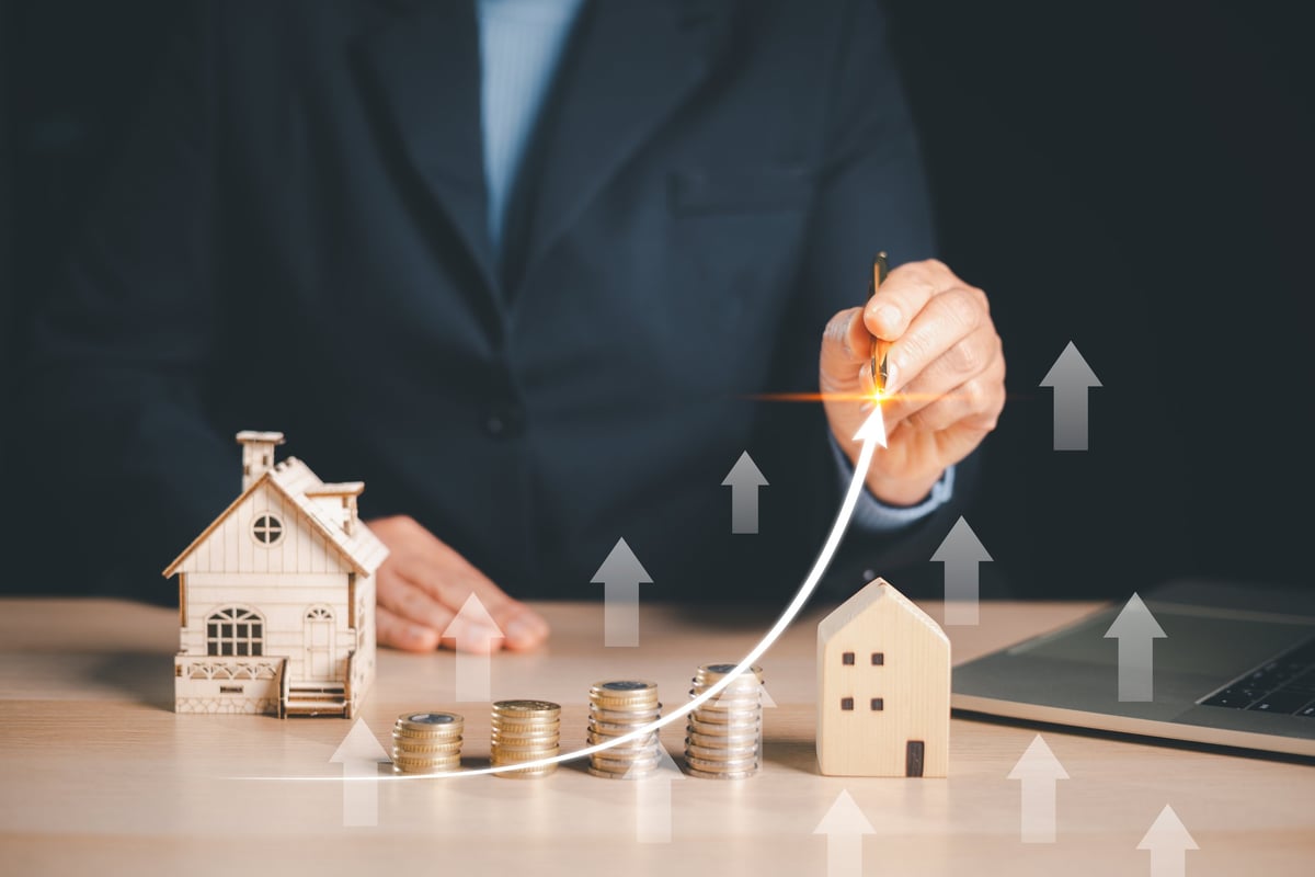 Businessman drawing rising arrow over coins and house models, showing real estate investment growth