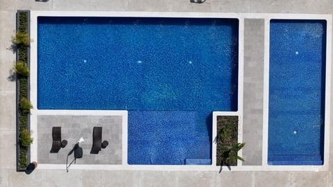 Aerial view of two connected swimming pools with loungers, surrounded by white concrete deck and green plants