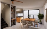 Modern open-concept apartment interior with dining table, wooden kitchen, stairs, and large windows overlooking greenery.
